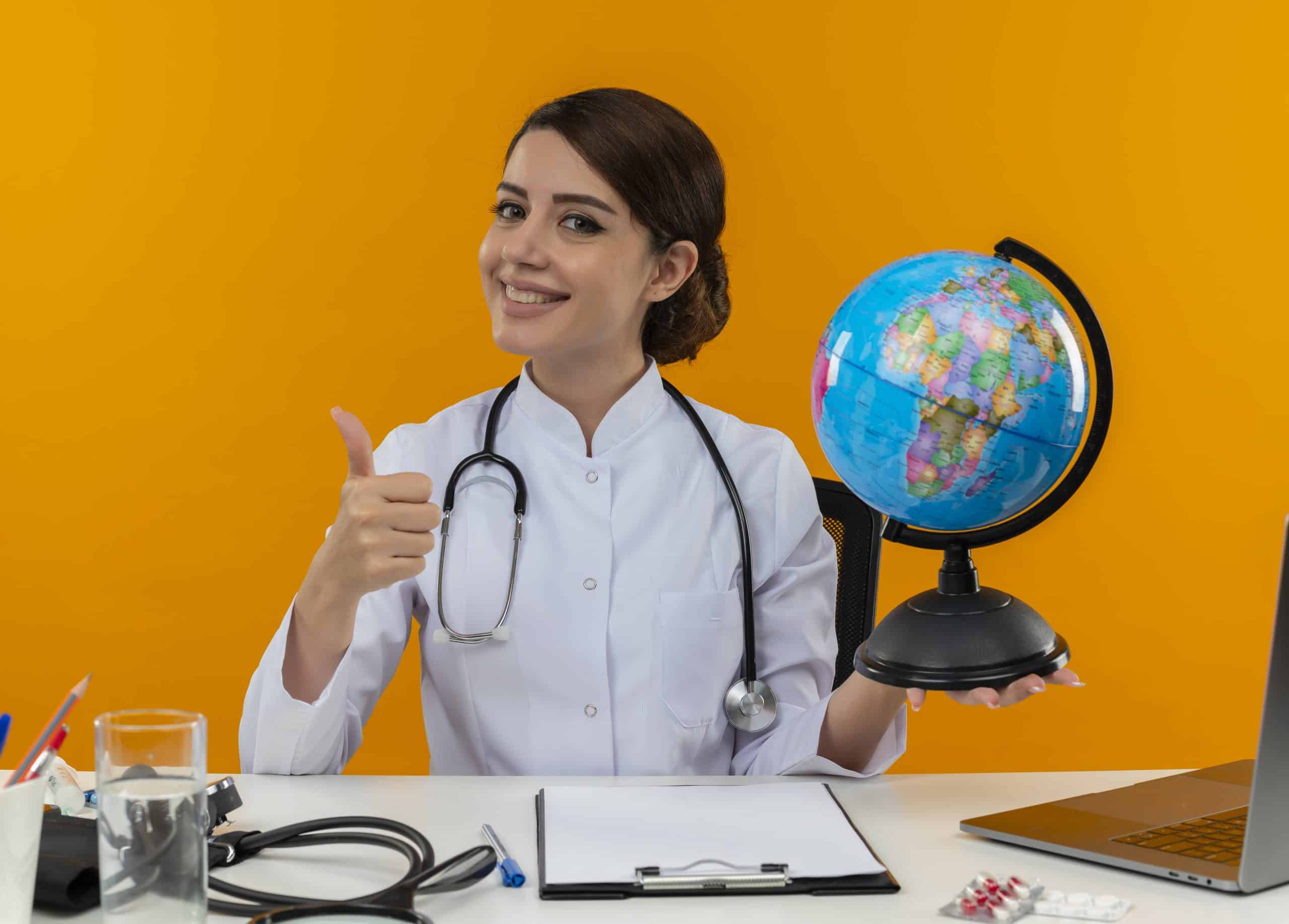 Médica sonriente trabajando en su ordenador con herramientas médicas y un globo terráqueo, signo de aprobación con el pulgar levantado.