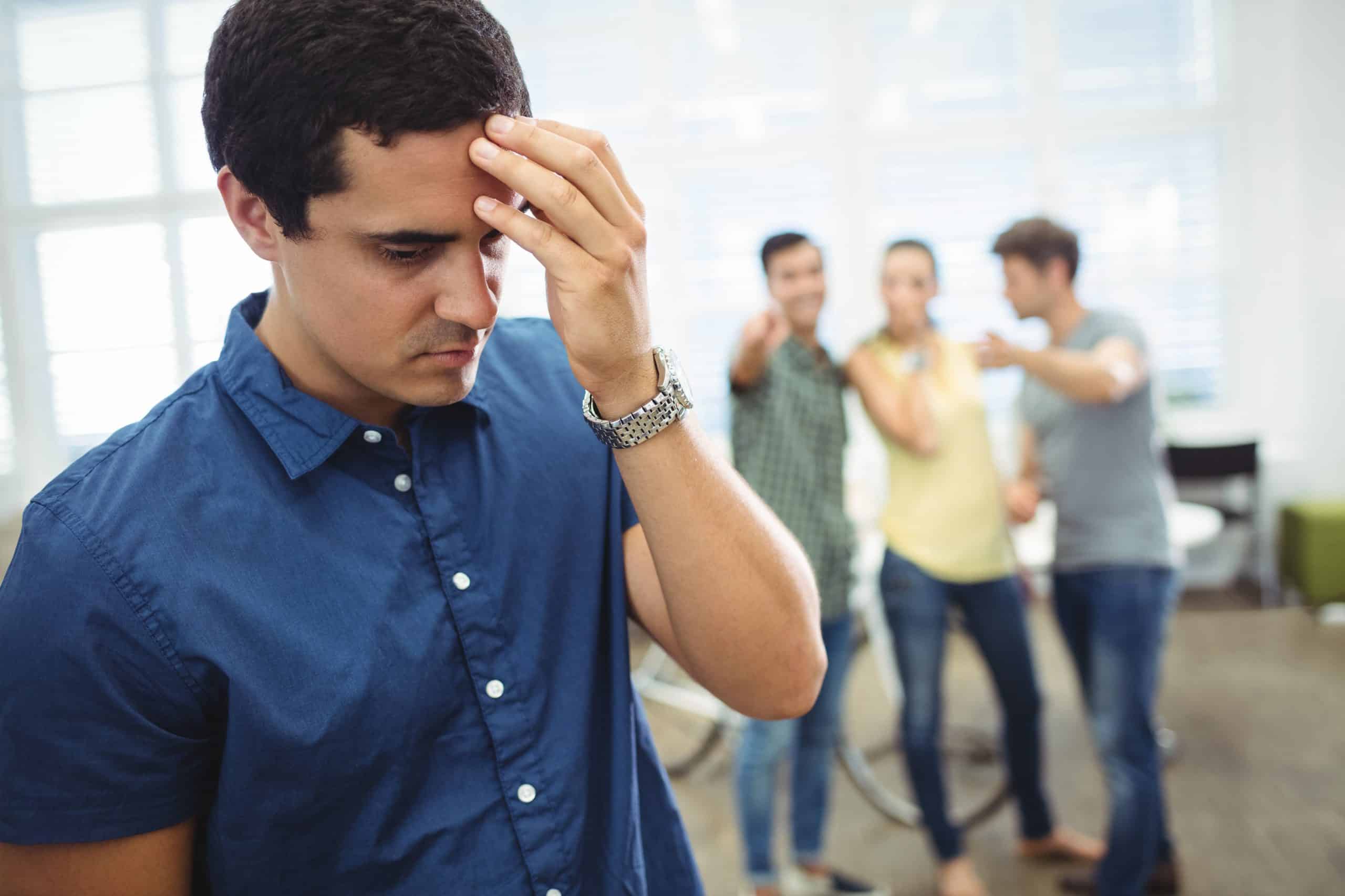 Hombre joven sintiendo presión laboral por no hablar inglés mientras compañeros cuchichean